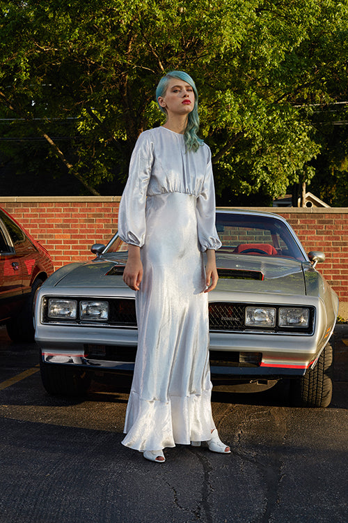 Cynthia Rowley 2019 Resort Collection features a long-sleeved silver, shimmery maxi dress that gathers at the high waist, and has a slight ruffle at the hem.