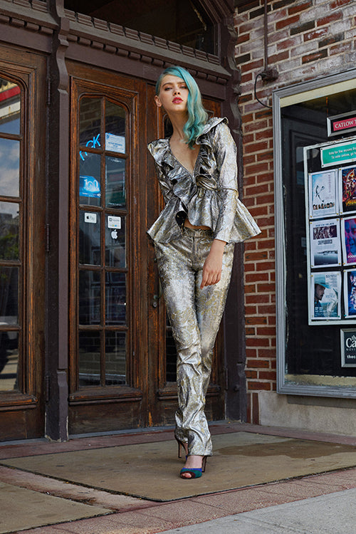 Cynthia Rowley 2019 Resort Collection features a metallic silver and gold patterned, long-sleeve ruffled top with a deep v-cut along with matching fitted pants, worn with blue and green heels. 