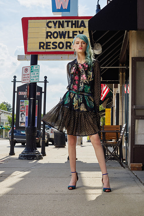 Cynthia Rowley Resort 2019 Collection features a black floral mini dress with a metallic finish paired with statement earrings, and two-toned, blue and green heels. 