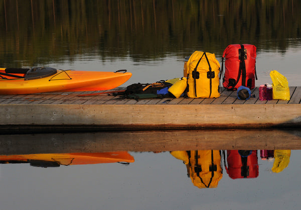 Packed up dry bags and backpacks on the dock beside a kayak on a river.