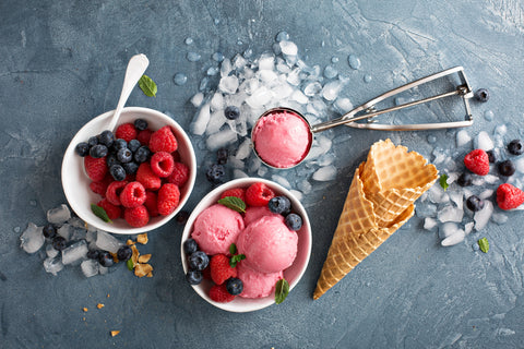Scoops of ice cream sit in a bowl on a table near an ice cream scoop, a bowl of berries, and waffle cones.