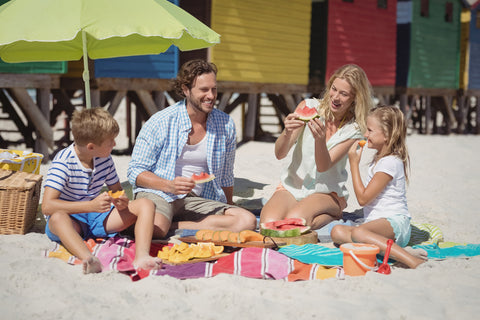 A family sits on a picnic blanket on the beach with a spread of foods laid out before them.
