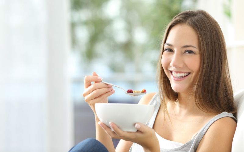 Frau mit braunem, langem Haar lacht in die Kamera. Sie hält eine weiße Schüssel in der einen und einen Löffel mit Müsli und Obst in der anderen Hand.