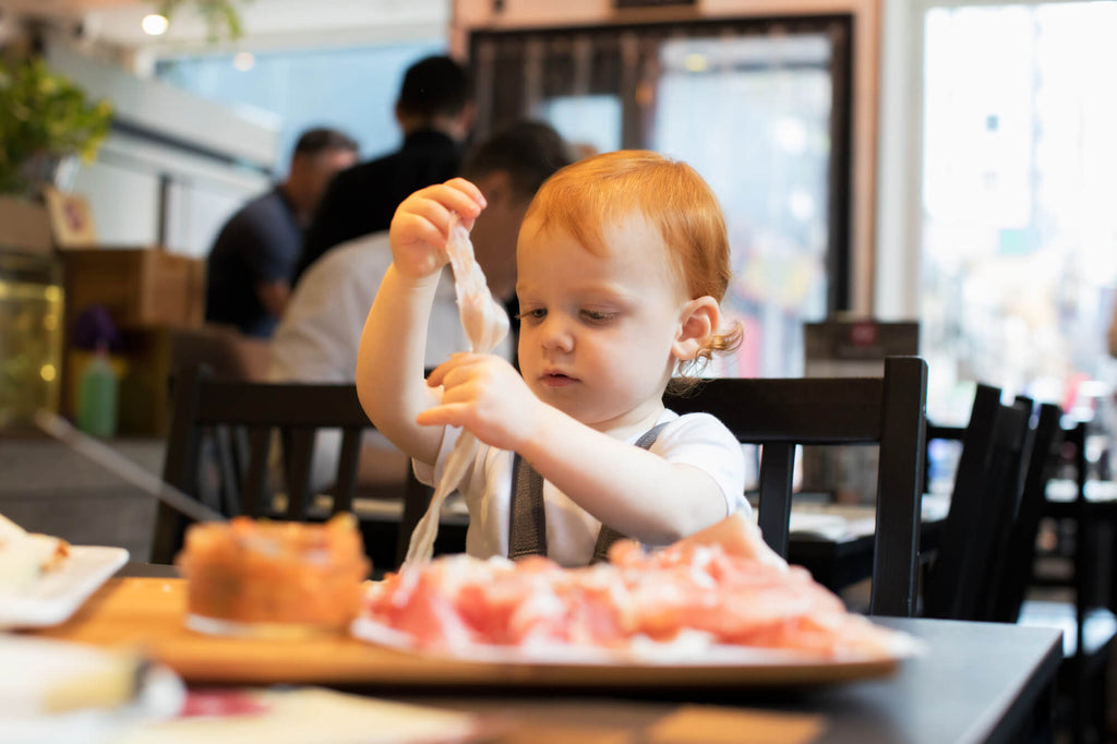 2 years old baby, sitting on a Bombol Foldable Booster Seat in a restaurant, playing with ham 