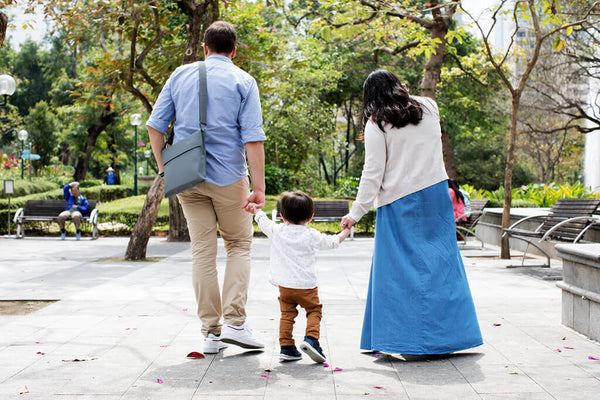 Parents holding baby’s hand walking in the park with foldable booster on father’s shoulder.