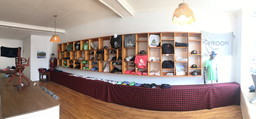 Cubbies on the wall of different shapes display Bark Eater Outfitters apparel Adirondack designs. A Buffalo red & black plaid camouflages a 2' high storage shelf. Items seen on the wall: Bark Eater unisex tee shirts, sweatshirts, mesh trucker hats