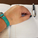 Psychology Professor Leslie Martinez' hand resting on a notebook next to pen. There is a gold transform mindful mark on left hand and a teal watch.