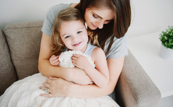 A mother lovingly hugging her young daughter as they sit on a couch, covered in a lush luxury Saranoni blanket.