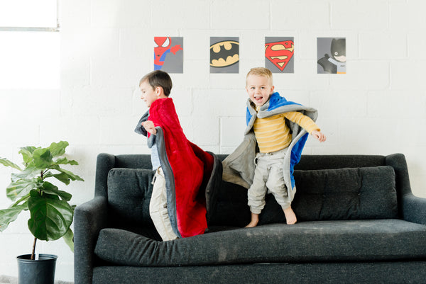 Two young friends jumping on the couch with their Saranoni Blankets tied on their shoulders like a cape.