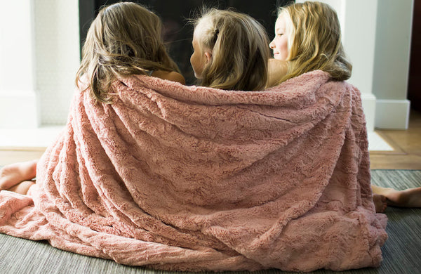 Three girls cuddle up near the fireplace in a super soft and fluffy Saranoni Luxury blanket.