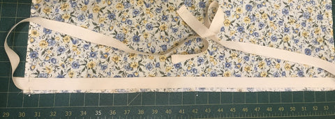 Along the bottom edge on the right side of the fabric, align the bottom of a 1 ½ yard piece of twill tape with the top of the serging and align the center of the tape with the center of the fabric.  Sew the bottom edge of the twill tape down 1/8” from the edge of the twill tape. Step 7
