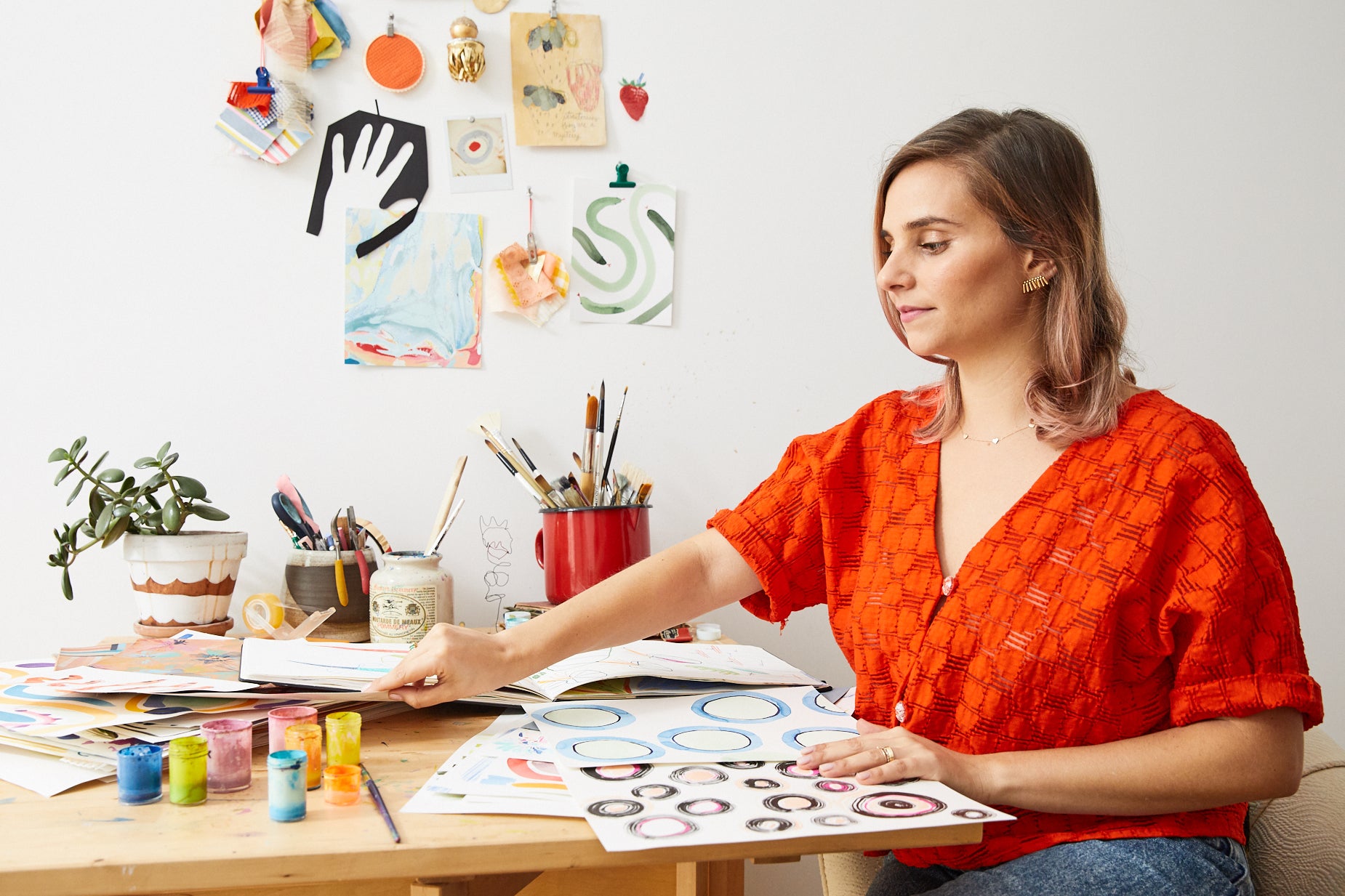 Brunette woman sitting at a desk with her watercolor artwork on the table and paint brush cans and whimsical artwork hanging on the wall