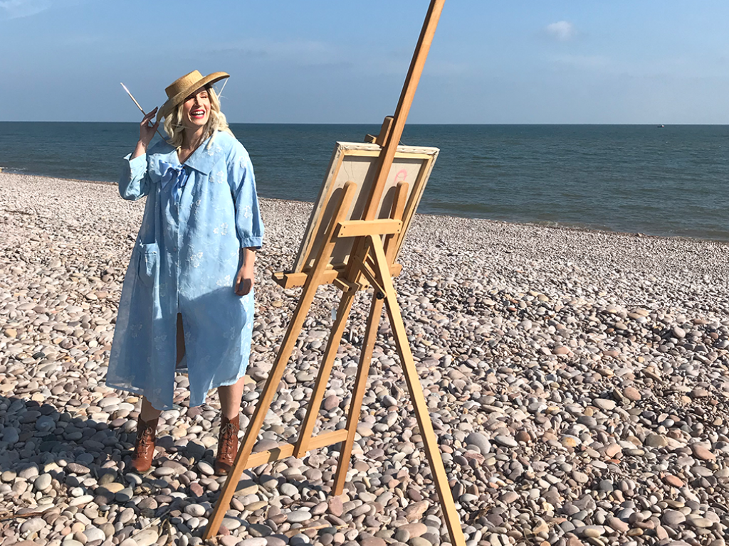 Influencer and blogger Jenny Mandera stands on a pebble beach on a bright sunny spring day. She is posed playfully at an easel, paintbrush in hand, and smiles into the middle distance, clutching her straw hat to her head against the sea breeze. She wears a distinctive blue swing coat  - an original vintage coat from the fifties.  