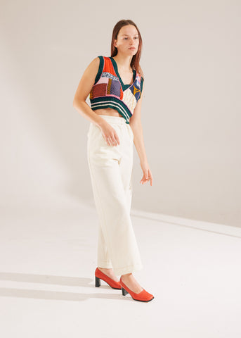Model poses in a studio setting with warm white lighting casting a shadow. Wearing cream semi tailored wide leg trousers and a hand knitted vest that is collaged into various sections. Coloured pieces include green ribbing with cream and pink highlights, cream cable knit, red cable knit and bright pink accents.