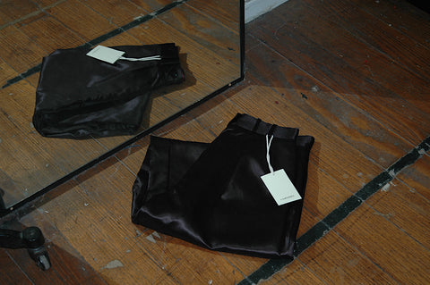 A pair of satin M.03 pants, folded and shot with its reflection in a nearby mirror. Shot in the Corepret studio on the ground, black pants contrast with the brown wooden floor boards
