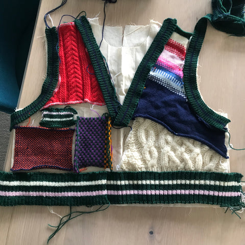 An image of a partially finished hand knitted vest. Deconstructed into each component featuring a red coloured cable knit section, cream cable knit piece, green ribbing with cream and pink highlights and a transitional shoulder piece that fades from lighter, pale pink colours right through to navy 