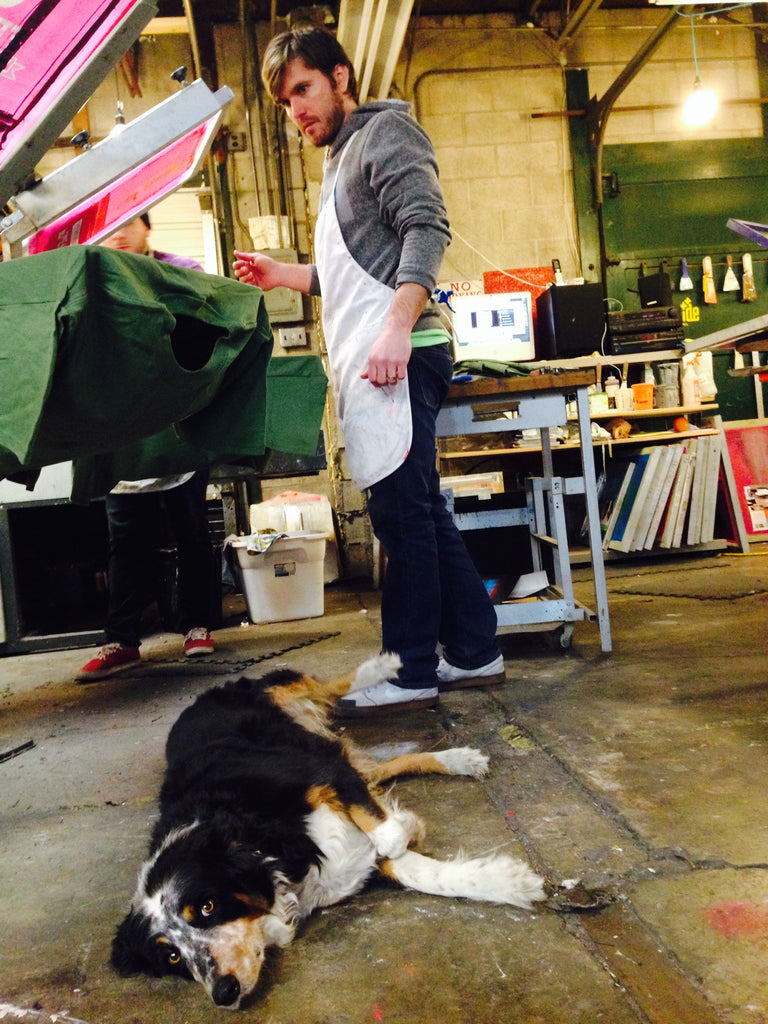 Mila on the floor of the screen printing shop with Nate at the machine.
