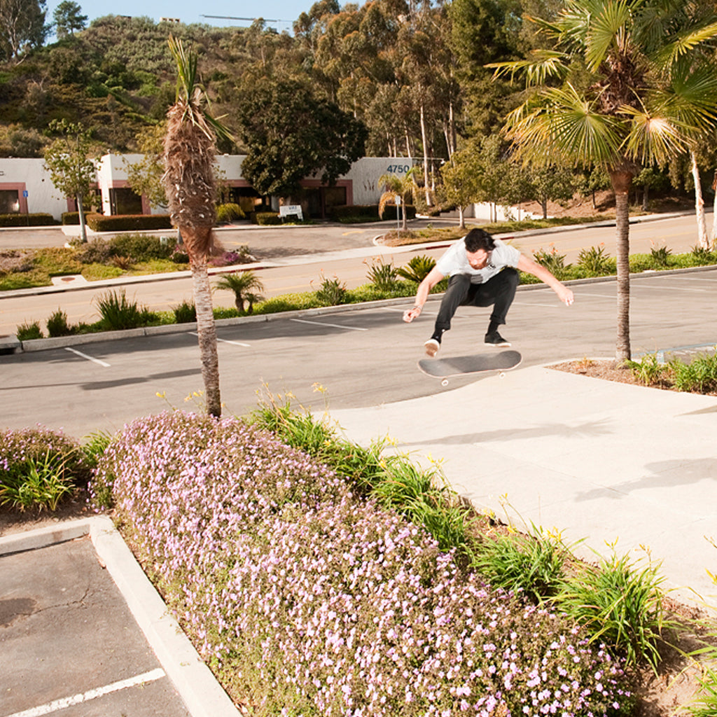 Corey Duffel doing a huge kickflip on his skateboard over a flower bed.