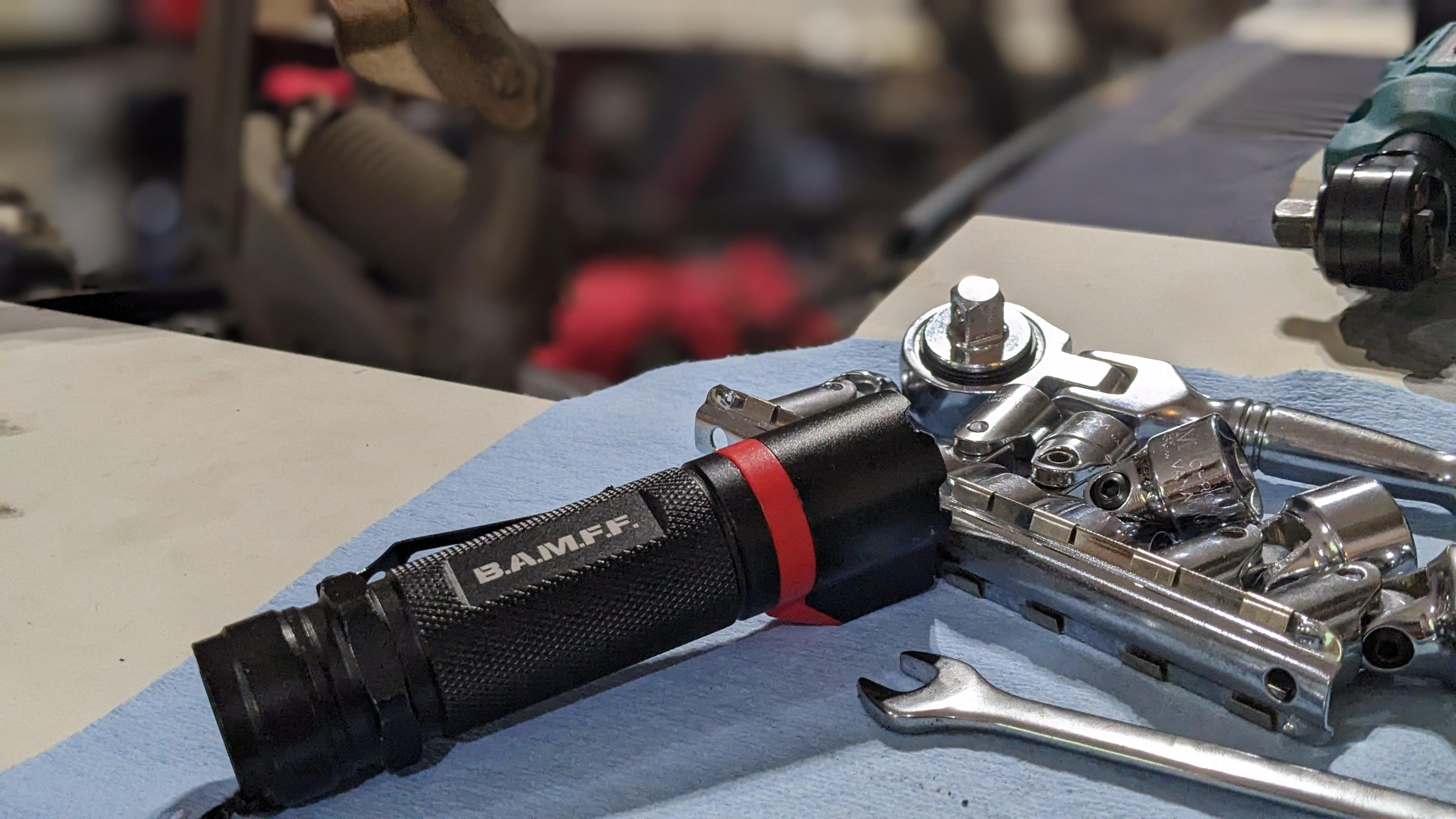BAMFF 8.0 Tactical Flashlight sitting on a blue shop towel with some sockets, socket wrench, and a box end wrench. Blurred out car engine bay in the background.