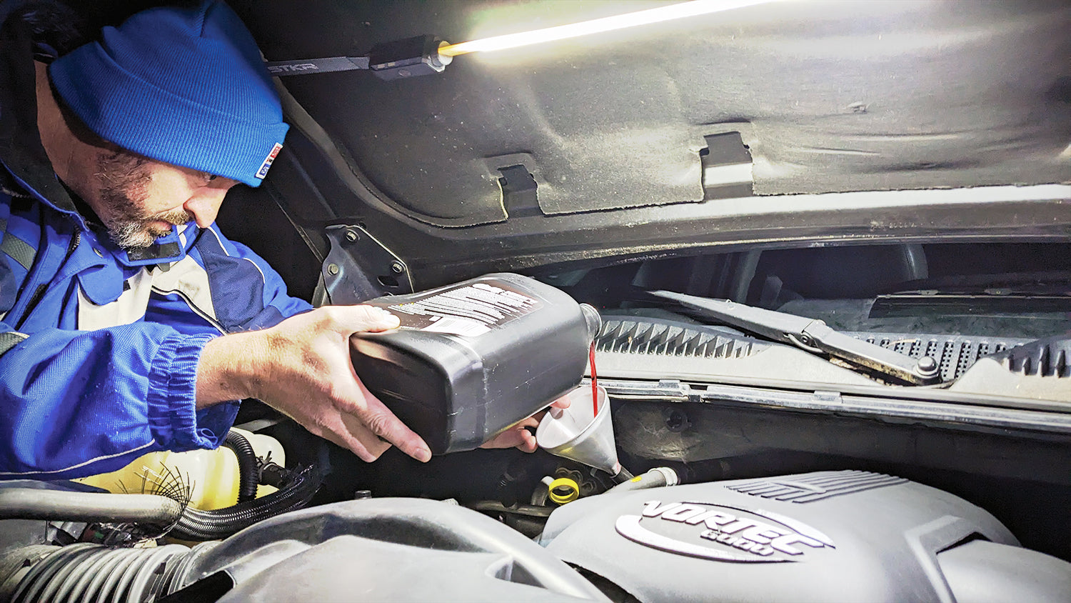 FLEXIT Under Hood Light use image showing a male pouring transmission fluid into a vehicle via a metal funnel. Night time scene light up by an Under Hood Light attached to the underside of the hood.