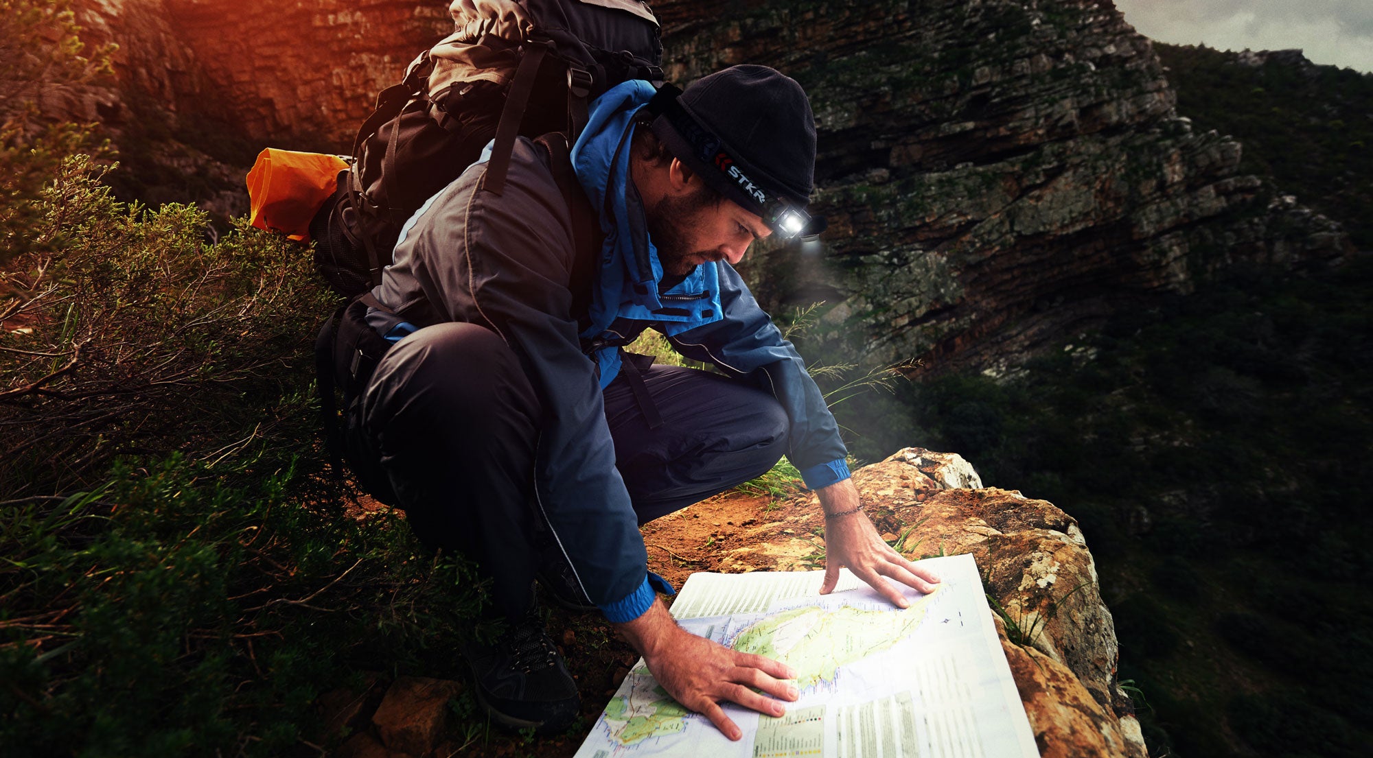 man hiking up in the mountains wearing an STKR headlamp kneeling down and looking at a map on the ground