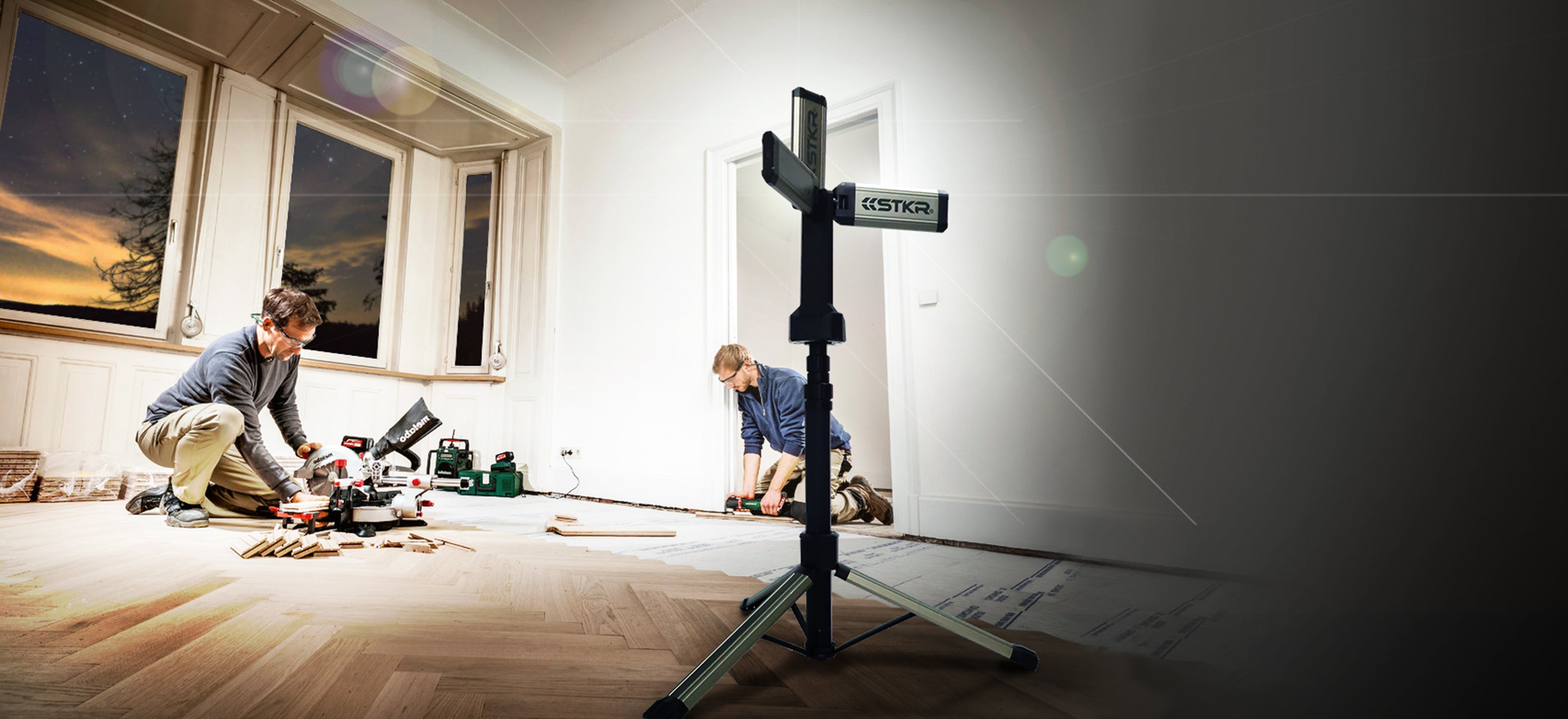 TRi-Mobile area work light on a tripod by STKR Concepts illuminating two male workers in the process of installing new hardwood flooring in a herringbone pattern. DIY home project after dark.