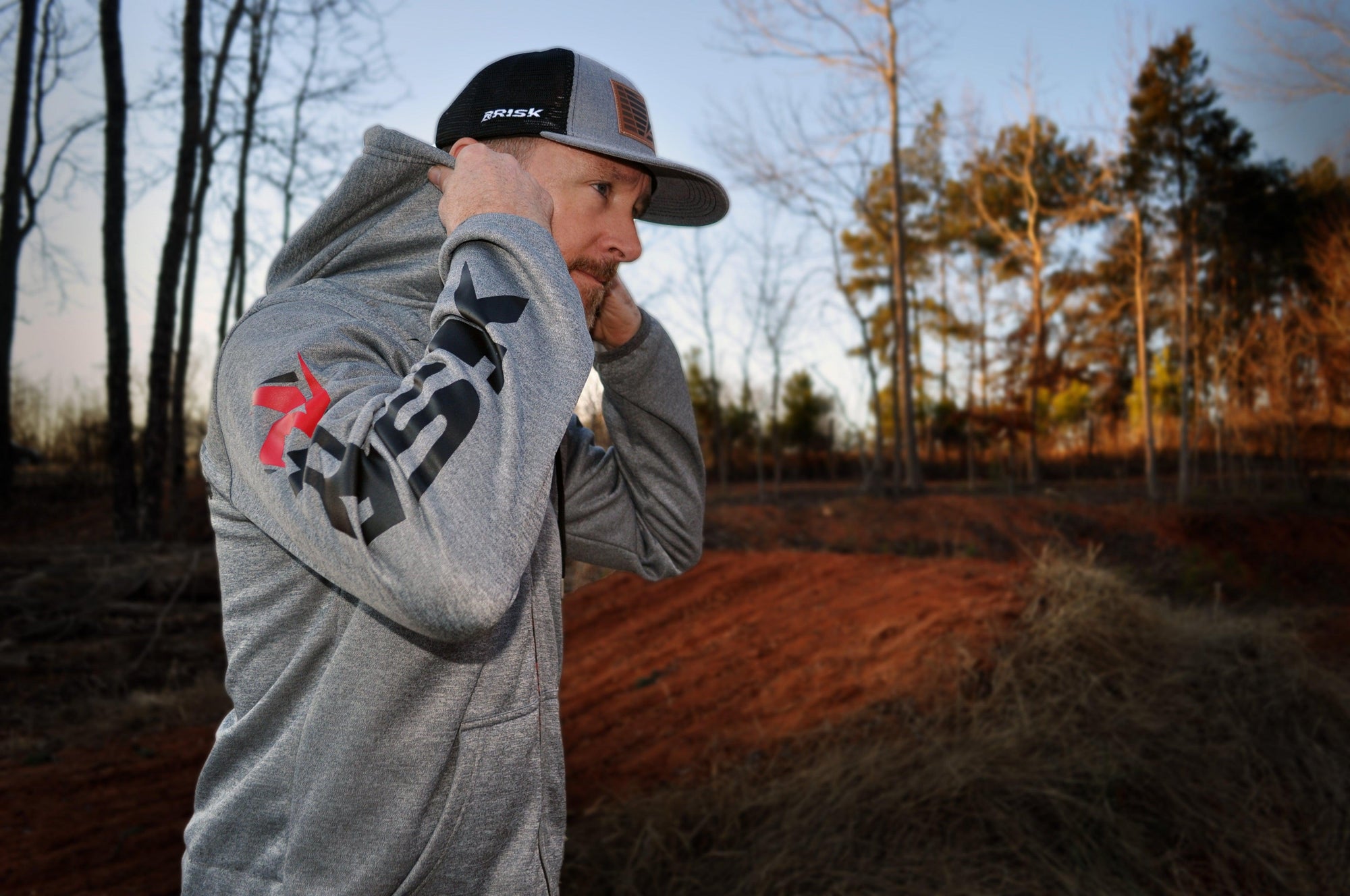 side view of a male wearing a Risk Racing zipper jacket pulling the hood onto his head. A dirt jump and tree scene blurred out in the background.