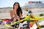 Risk Racing's March Moto Model Amber Juliana wearing a white bikini standing behind a motocross bike resting her forearms on the seat - medium shot - white fenced off MX track in background
