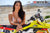 Risk Racing's March Moto Model Amber Juliana wearing a bikini standing behind a motocross bike resting her forearms on the seat - medium shot - white fenced off MX track in background