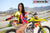 Risk Racing's March Moto Model Amber Juliana wearing a Yellow and Red VENTilate V2 Jersey and white white bikini bottoms standing three quarter pose in front of a motocross bike sitting on an ATS stand - medium shot - white fenced off MX track in background