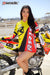 Risk Racing's March Moto Model Amber Juliana wearing a Yellow and Red VENTilate V2 Jersey that's covering any sign of her white bikini bottoms in front of a motocross bike sitting on an ATS stand - medium shot - white fenced off MX track in background