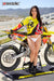 Risk Racing's March Moto Model Amber Juliana wearing a Yellow and Red VENTilate V2 Jersey and white bikini bottoms in front of a motocross bike sitting on an ATS stand - medium shot - white fenced off MX track in background
