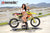 Risk Racing's March Moto Model Amber Juliana wearing a white bikini in front of a motocross bike that's sitting on an ATS Stand- wide shot - white fenced off MX track in background