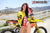 Risk Racing's March Moto Model Amber Juliana tugging on a Yellow and Red VENTilate V2 Jersey in front of a motocross bike - close up shot - white fenced off MX track in background