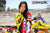 Risk Racing's March Moto Model Amber Juliana wearing a Yellow and Red VENTilate V2 Jersey with one hand up on her neck and the other pulling the Jersey down making it appear that she has no bottoms on while posing in front of a motocross bike - close up shot - white fenced off MX track in background