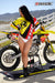 Risk Racing's March Moto Model Amber Juliana wearing a Yellow and Red VENTilate V2 Jersey and white bikini bottoms with her hands on her hips looking off screen left in front of a motocross bike that's sitting on an ATS stand - medium shot - white fenced off MX track in background