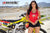 Risk Racing's March Moto Model Amber Juliana wearing a red Risky moto chick daisy tank top and black short shorts with both hands near her waist while posing in front of a motocross bike - medium shot - white fenced off MX track in background