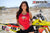 Risk Racing's March Moto Model Amber Juliana wearing a red Risky moto chick daisy tank top and black short shorts with both hands near her waist while posing in front of a motocross bike - close up shot - white fenced off MX track in background