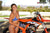 Risk Racing's May Moto Model Natalie Nicole wearing a 2 piece bikini posing in front of a motocross bike. Both forearms near her mid-drift hands lightly crossed. - close up shot - white fenced off MX track in background