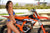 Risk Racing's May Moto Model Natalie Nicole wearing a 2 piece bikini posing sideways in front of a motocross bike. Her buns are both showing as she arches back and looks over her right shoulder. pose 3 - close up shot - white fenced off MX track in background
