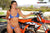 Risk Racing's May Moto Model Natalie Nicole wearing a 2 piece bikini posing in front of a motocross bike. Left hand on the throttle and right lightly touching her bikini bottom strap. - close up shot - white fenced off MX track in background