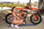 Risk Racing's May Moto Model Natalie Nicole wearing a 2 piece bikini kneeling sideways in front of a motocross bike that's sitting on an ATS Stand and Factory Pit mat by Risk Racing. Her left hand on the seat and right resting on her knee. Turning her buns towards camera - pose 2 - wide shot - white fenced off MX track in background