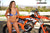 Risk Racing's May Moto Model Natalie Nicole wearing a 2 piece bikini posing in front of a motocross bike that's sitting on an ATS Stand by Risk Racing. Both her hands are slightly pulling up on her bikini bottom straps. - medium close shot - white fenced off MX track in background
