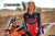 June Moto Model Rochelle Roche wearing a Risk Racing Factory Pit Dry-Fit Shirt. She's standing in front of an orange dirt bike. MX track in the background. Pose #3