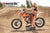 June Moto Model Rochelle Roche wearing a 2 piece black bikini in front of an orange dirt bike with her right hand on the handlebar grip. Bike is sitting on an Adjustable Top Stand by Risk Racing. MX track in the background.
