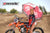 June Moto Model Rochelle Roche wearing a Risk Racing Factory Pit Dry-Fit Shirt and holding a factory pit umbrella. She's standing in front of an orange dirt bike. MX track in the background.