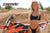 Risk Racing's June Moto Model Rochelle Roche wearing a 2 piece black bikini posing in front of an orange dirt bike with her right hand on the gas tank, left hand on the rear fender. MX track in the background.