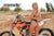 Risk Racing's June Moto Model Rochelle Roche wearing a 2 piece black bikini posing backwards, buns facing camera, in front of an orange dirt bike. She's looking back at us over her left shoulder with an MX track in the background.