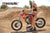 June Moto Model Rochelle Roche wearing a 2 piece black bikini in front of an orange dirt bike with her right hand on the handlebar grip. Bike is sitting on an Adjustable Top Stand by Risk Racing. MX track in the background.