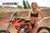 Risk Racing's June Moto Model Rochelle Roche wearing a 2 piece black bikini in front of an orange dirt bike with her right hand on the gas tank, left hand pulling up on her bikini bottom strap. MX track in the background.
